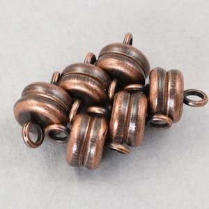 May include: A collection of copper-colored magnetic clasps. Each clasp has a cylindrical shape with a ridged design and a small loop at each end. The clasps are clustered together on a light gray surface.