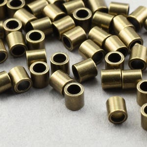 May include: A collection of small, cylindrical brass beads with a hole running through the center. These beads are scattered across a light gray surface, showcasing their metallic sheen and simple design. Ideal for jewelry making.