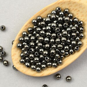 May include: A collection of small, dark gray metallic beads, with a shiny finish, are piled in a wooden scoop. Some beads are scattered around the scoop on a light gray surface. These beads are likely used for jewelry making or crafting.