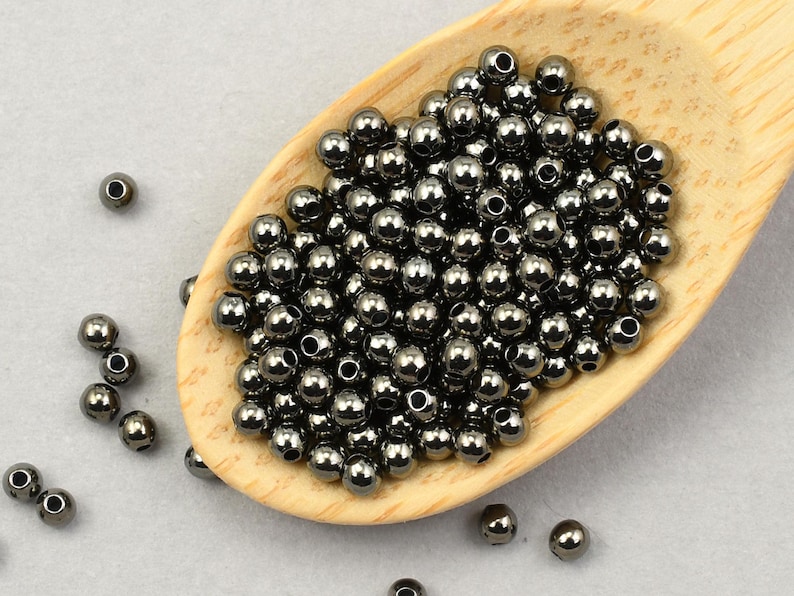 May include: A collection of small, dark gray metallic beads, with a shiny finish, are piled in a wooden scoop. Some beads are scattered around the scoop on a light gray surface. These beads are likely used for jewelry making or crafting.