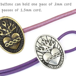 May include: Two oval-shaped metal buttons with a heart and flame design. The button on the left is gold-toned and the button on the right is silver-toned.  The text on the image reads: "These buttons can hold one pass of 2mm cord or two passes of 1.5mm cord."