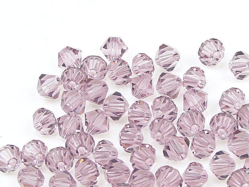 May include: A scattering of light purple glass beads with a faceted, star-shaped design. The beads are arranged on a white background.