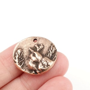 May include: A round copper charm with a detailed relief of a bird perched on a branch. The charm has a hole at the top for attaching to a necklace or bracelet.