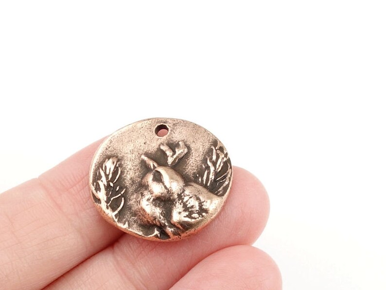 May include: A round copper charm with a detailed relief of a bird perched on a branch. The charm has a hole at the top for attaching to a necklace or bracelet.