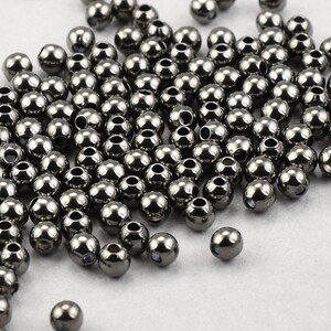 May include: A collection of small, metallic gray beads with a shiny finish. Each bead has a hole running through the center, suggesting they are for stringing or crafting. The beads are scattered across a light-colored surface.