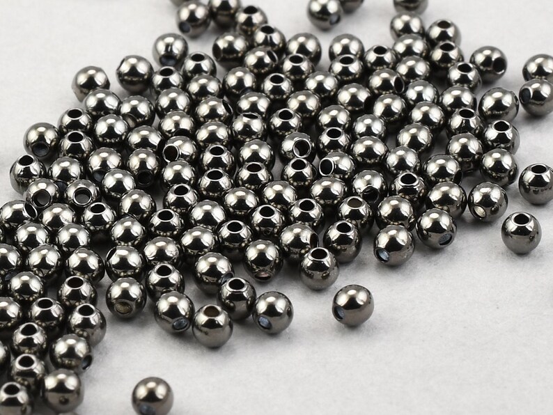 May include: A collection of small, metallic gray beads with a shiny finish. Each bead has a hole running through the center, suggesting they are for stringing or crafting. The beads are scattered across a light-colored surface.