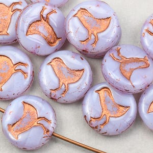 May include: A collection of round, lavender glass beads with a copper-colored bird design. The beads are approximately 1 inch in diameter.
