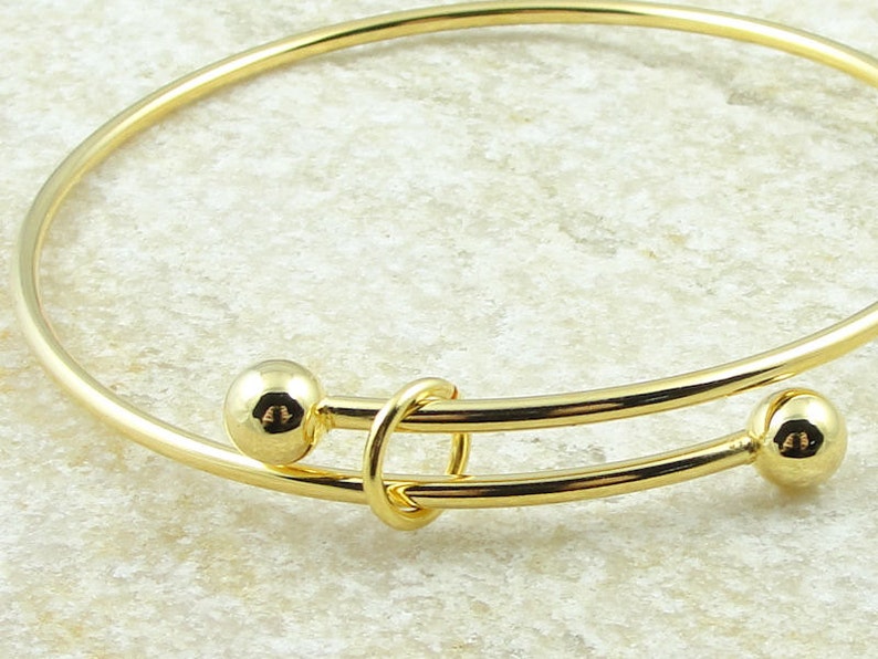 May include: A gold-colored bangle bracelet with two small, round, gold-colored beads on either side of a decorative clasp. The bracelet is made of a thin, curved metal band.