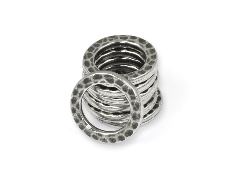 May include: A stack of silver-colored metal rings with a hammered texture. The rings are circular with a slightly irregular surface, creating a rustic or industrial aesthetic. The rings are stacked on a white background.