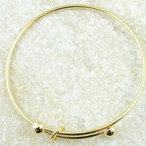 May include: A gold-colored bangle bracelet with a simple, elegant design. The bracelet features a circular band with a decorative clasp and two small spherical accents. The metal has a polished finish, reflecting light.