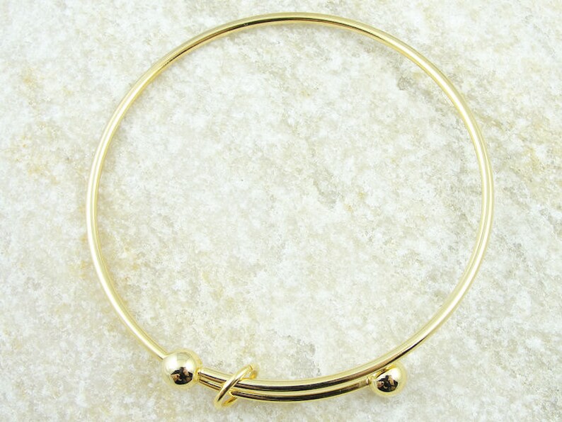 May include: A gold-colored bangle bracelet with a simple, elegant design. The bracelet features a circular band with a decorative clasp and two small spherical accents. The metal has a polished finish, reflecting light.
