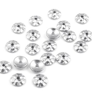 May include: Silver-colored metal jewelry components with a hammered texture. These small, round components have a central hole and a slightly concave back. The components are scattered on a white surface.