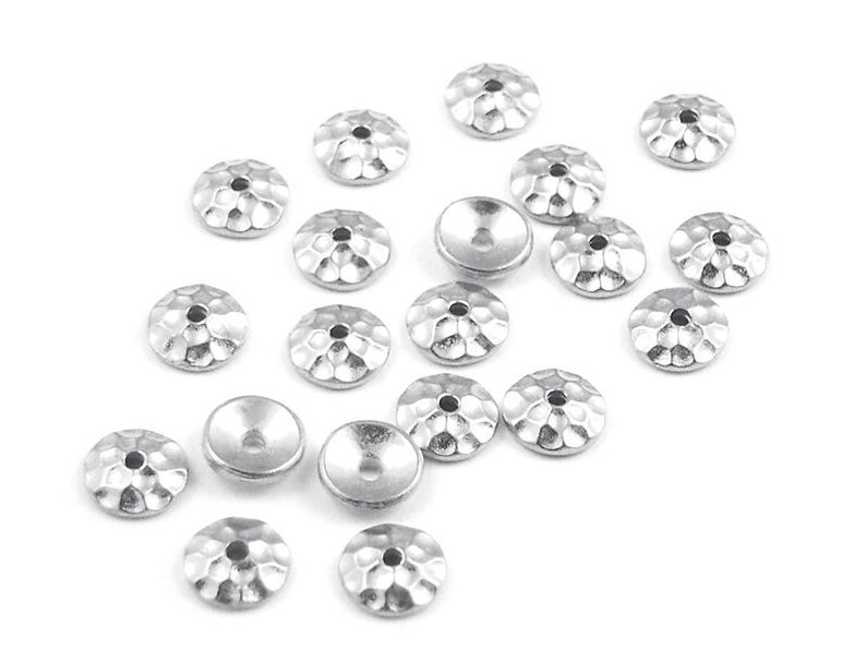 May include: Silver-colored metal jewelry components with a hammered texture. These small, round components have a central hole and a slightly concave back. The components are scattered on a white surface.