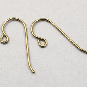 Puede incluir: Dos ganchos dorados para pendientes se muestran sobre un fondo claro. Los ganchos están hechos de alambre de metal fino y curvado, con un diseño en bucle en la parte superior y una parte inferior recta ligeramente curvada.