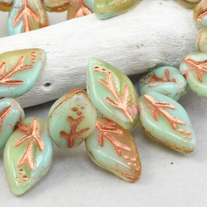 May include: A strand of light green glass beads with copper leaf designs. The beads are shaped like leaves and have a slightly iridescent finish.