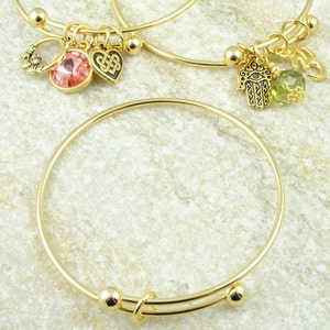 May include: Gold-toned adjustable bangle bracelets. One bracelet features a pink gemstone and a Celtic knot charm. Another has a green gemstone and a Hamsa hand charm. The bracelets are displayed on a light-colored, textured surface.