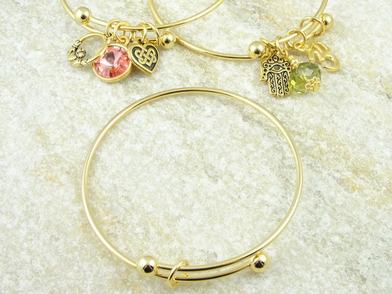 May include: Gold-toned adjustable bangle bracelets. One bracelet features a pink gemstone and a Celtic knot charm. Another has a green gemstone and a Hamsa hand charm. The bracelets are displayed on a light-colored, textured surface.