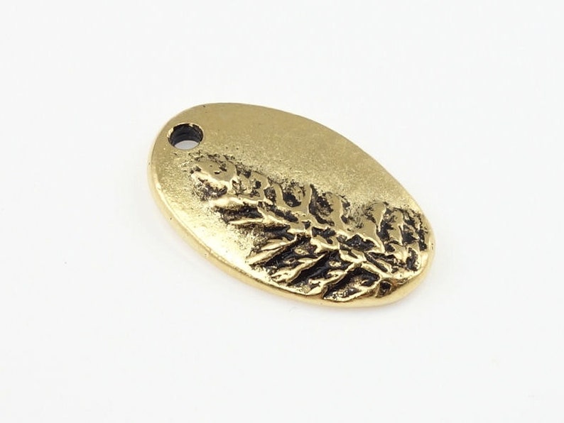 May include: Gold-toned oval charm with a raised design of a sprig of leaves. The charm has a small hole at the top for attaching to a necklace or bracelet.