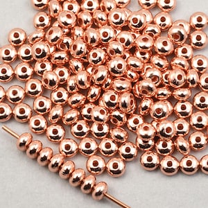 May include: A large pile of rose gold colored round beads. The beads have a slightly textured surface and are scattered on a white background. A single strand of the beads is strung on a thin wire.
