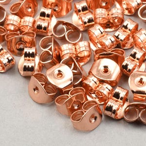 May include: A collection of shiny, copper-colored earring backs. The earring backs are in various shapes and sizes, with a smooth, reflective surface. The image is a close-up shot, showing the details of the jewelry findings.