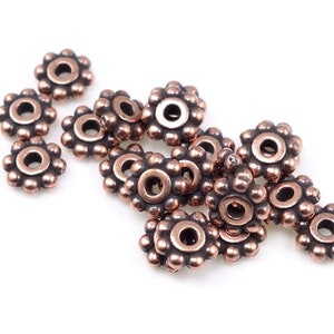May include: A group of 14 copper-coloured metal beads with a flower-shaped design. Each bead has a small hole in the centre.