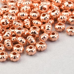 May include: A collection of rose gold-colored metal beads. The beads have a flower-like shape with multiple petals and a hole through the center. The beads are scattered on a light gray surface.