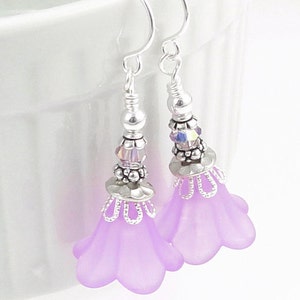 May include: A pair of lavender flower earrings with silver-colored accents. The earrings feature a delicate design with small beads and a floral shape. The hooks are silver-colored. The earrings are designed to be worn as a fashion accessory.