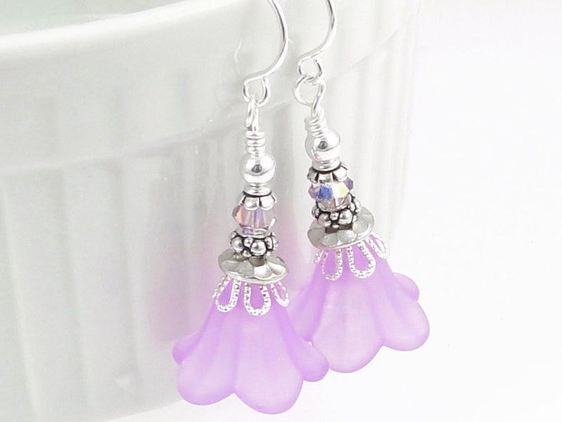 May include: A pair of lavender flower earrings with silver-colored accents. The earrings feature a delicate design with small beads and a floral shape. The hooks are silver-colored. The earrings are designed to be worn as a fashion accessory.