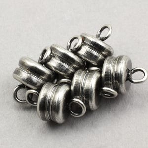 May include: A collection of silver-toned magnetic clasps. Each clasp has a cylindrical shape with a grooved design and a small loop for attaching to jewelry. The clasps are clustered together, showcasing their metallic finish.