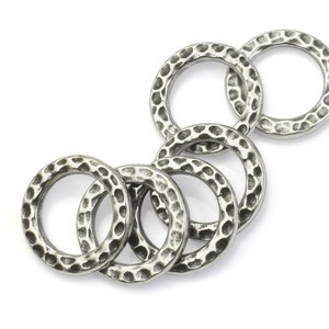 May include: A collection of six silver-colored metal rings with a hammered texture. The rings are circular and have a slightly irregular surface, creating a rustic appearance. The rings are arranged in a loose, overlapping pattern against a white background.