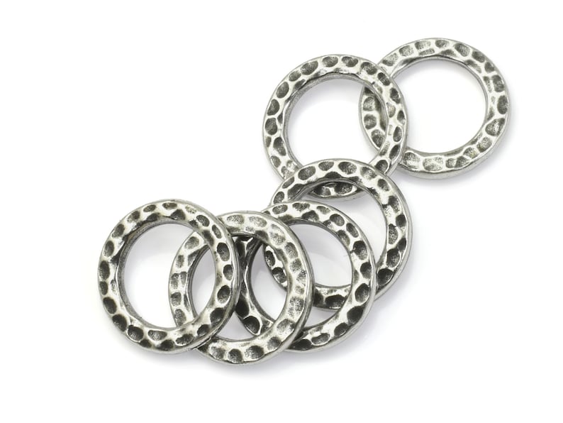 May include: A collection of six silver-colored metal rings with a hammered texture. The rings are circular and have a slightly irregular surface, creating a rustic appearance. The rings are arranged in a loose, overlapping pattern against a white background.