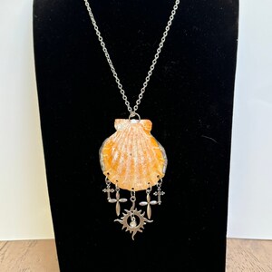 May include: A silver chain necklace with a large seashell pendant. The pendant has a silver sun charm with a small rhinestone in the center and five silver cross charms hanging from it.