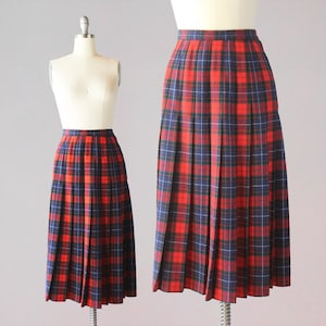 May include: A vintage pleated skirt in a red, navy, and white tartan pattern. The skirt is displayed on a mannequin, showing its full length and fabric details. The skirt has a classic design, suitable for various occasions.