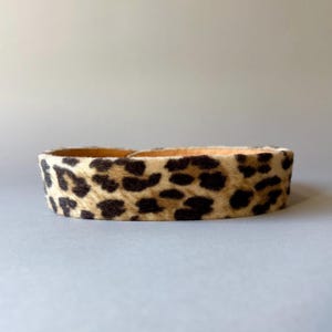Puede incluir: Un brazalete ancho con estampado de leopardo. El brazalete presenta una base beige con manchas marrones oscuras. El interior del brazalete es de color marrón claro. Probablemente está hecho de piel de animal o material similar.