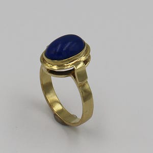 Könnte beinhalten: Ein goldener Ring mit einem großen, ovalen, tiefblauen Edelstein. Der Edelstein ist in eine goldene Lünette gefasst, und der Ring hat ein schlichtes, elegantes Design. Der Ring besteht wahrscheinlich aus Gold und einem Lapislazuli-Edelstein.