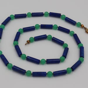 Könnte beinhalten: Halskette mit abwechselnd dunkelblauen zylindrischen Perlen und kleinen grünen kugelförmigen Perlen. Die Halskette hat einen goldfarbenen Verschluss. Die Perlen sind in einem kreisförmigen Muster auf weißem Hintergrund angeordnet.