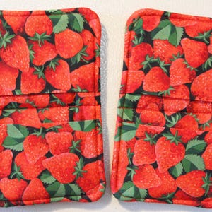 May include: Two rectangular fabric pot holders featuring a repeating pattern of red strawberries and green leaves on a black background. Each item has a horizontal stitched pocket across the center and is finished with matching red fabric binding.