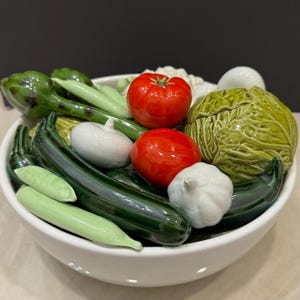 Puede incluir: Un cuenco de cerámica blanco lleno de verduras de cerámica. Las verduras incluyen calabacín verde, tomates, ajo, alcachofas y una col. Las verduras son de colores brillantes y están dispuestas de forma decorativa.