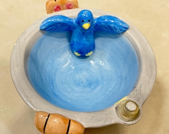 Könnte beinhalten: Ein Keramik-Aschenbecher mit einer blauen Vogelfigur in der Mitte. Der Aschenbecher hat einen hellblauen Innenraum und einen grauen Rand. Dekorative Elemente sind ein kleines, hellbraunes Brotstück und eine kleine, goldfarbene Glocke.