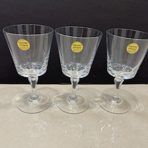May include: Three clear stemmed wine glasses with a flared bowl and a round base. Each glass has a yellow sticker that reads "Cristal d'Arques". The glasses are made of clear glass and are displayed on a light-colored surface.