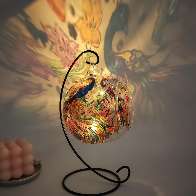 K&ouml;nnte beinhalten: Eine Glaskugel mit einem schwarzen Metallst&auml;nder. Die Kugel ist mit einem bunten Pfauenmuster verziert und verf&uuml;gt &uuml;ber ein Licht im Inneren. Die Kugel wirft einen Schatten des Pfauenmusters an die Wand hinter ihr.
