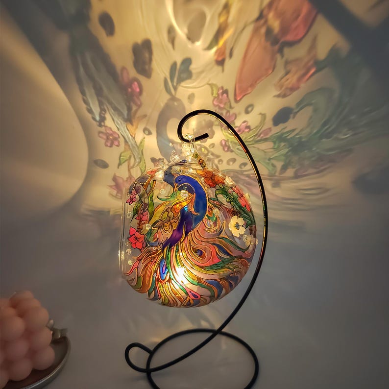 K&ouml;nnte beinhalten: Eine Glaskugel mit einem schwarzen Metallst&auml;nder, die mit einem handgemalten Pfauendesign in leuchtenden Farben verziert ist, darunter Blau, Gr&uuml;n, Pink und Gelb.