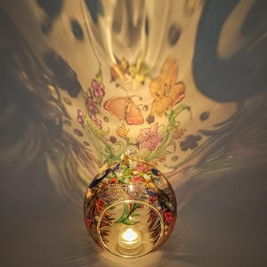 K&ouml;nnte beinhalten: Ein runder Glas-Kerzenhalter mit einem darauf gemalten Blumen- und Schmetterlingsmuster. Der Kerzenhalter ist mit einem Teelicht im Inneren beleuchtet.