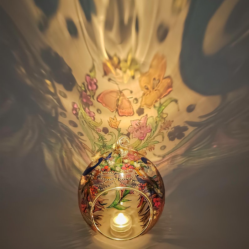K&ouml;nnte beinhalten: Ein runder Glas-Kerzenhalter mit einem darauf gemalten Blumen- und Schmetterlingsmuster. Der Kerzenhalter ist mit einem Teelicht im Inneren beleuchtet.