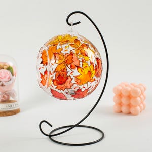 Puede incluir: Un portavelas de vidrio con un soporte de metal negro. El vidrio está decorado con hojas de otoño pintadas en tonos naranja, amarillo y rojo.