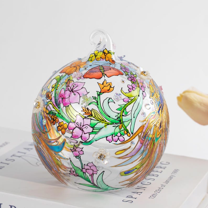 K&ouml;nnte beinhalten: Ein transparenter Glaskugelschmuck mit handgemaltem Design aus Blumen, Bl&auml;ttern und einem Schmetterling. Der Kugelschmuck ist mit goldenen Akzenten und schimmerndem Glitzer verziert.