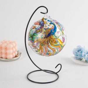 K&ouml;nnte beinhalten: Ein gl&auml;serner Weihnachtsbaumschmuck mit einem Pfauenmotiv, das in leuchtenden Farben wie Blau, Gold und Rosa bemalt ist. Der Schmuck wird auf einem schwarzen Metallst&auml;nder pr&auml;sentiert.