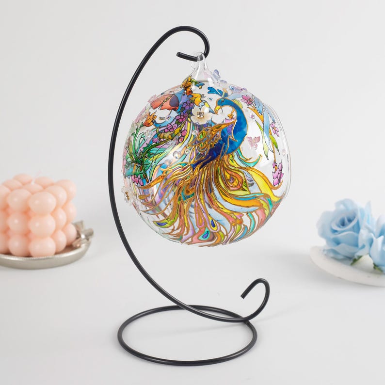 K&ouml;nnte beinhalten: Ein gl&auml;serner Weihnachtsbaumschmuck mit einem Pfauenmotiv, das in leuchtenden Farben wie Blau, Gold und Rosa bemalt ist. Der Schmuck wird auf einem schwarzen Metallst&auml;nder pr&auml;sentiert.