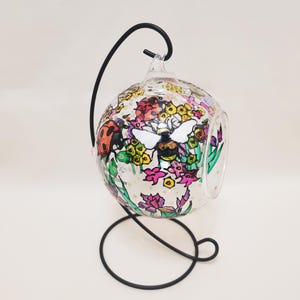 May include: A clear glass terrarium with a black metal stand. The terrarium is decorated with colorful flowers, a ladybug, and a bee. The stand has a circular base and a curved top.