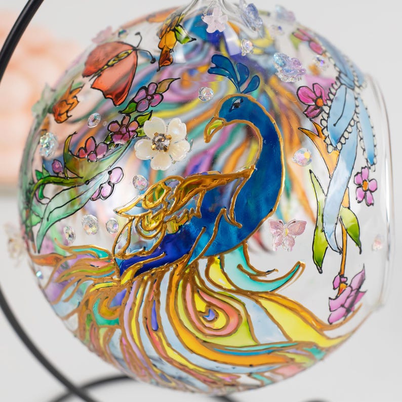 K&ouml;nnte beinhalten: Ein Glasornament mit handbemaltem Pfauendesign. Der Pfau ist blau mit goldenen Akzenten und einem bunten Schwanz. Das Ornament ist mit Blumen, Schmetterlingen und Strasssteinen verziert.